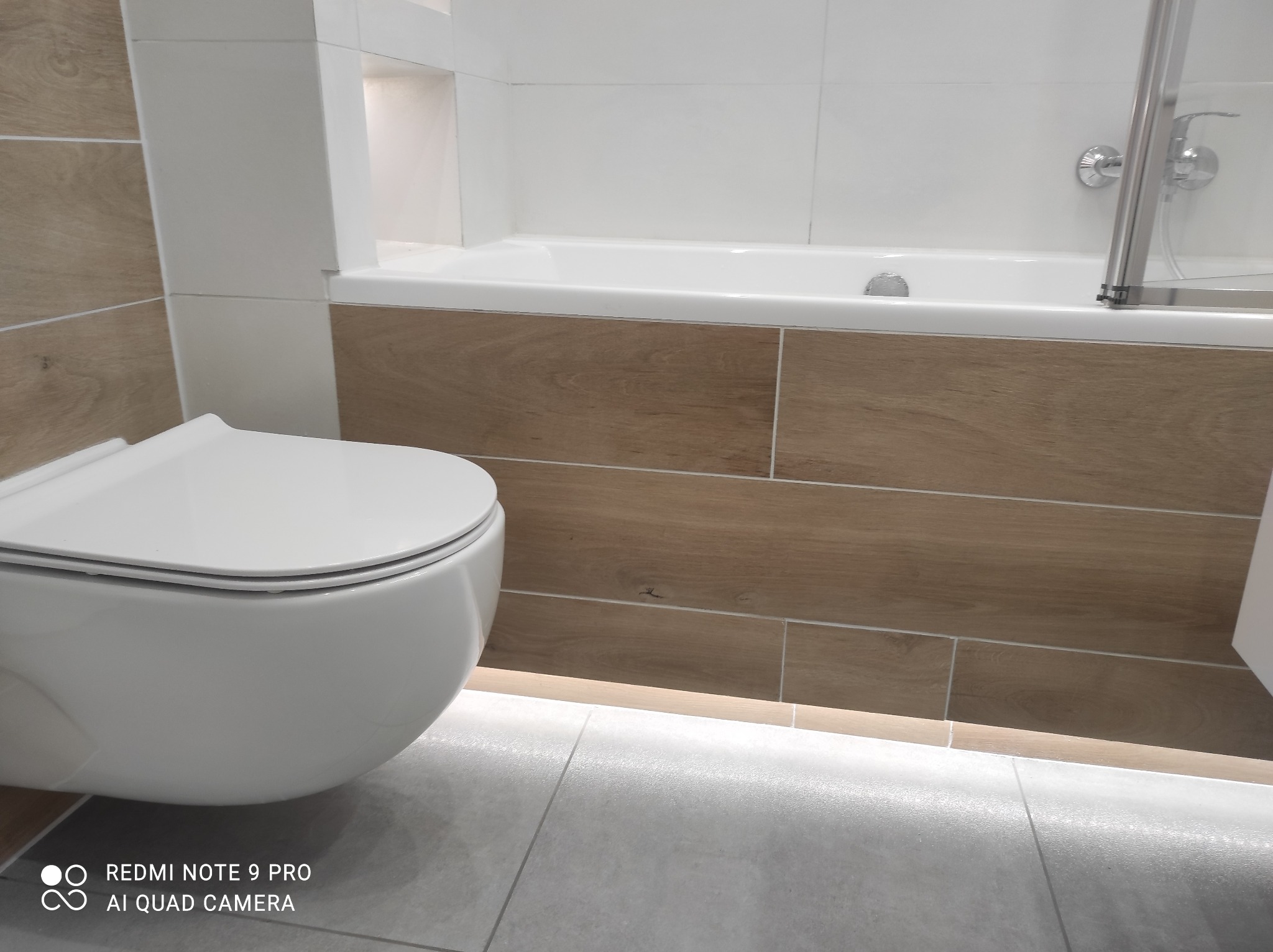 Nowoczesna, minimalistyczna łazienka z podwieszaną toaletą, obudową wanny imitującą drewno i szarą podłogą z delikatnym oświetleniem LED pod wanną.