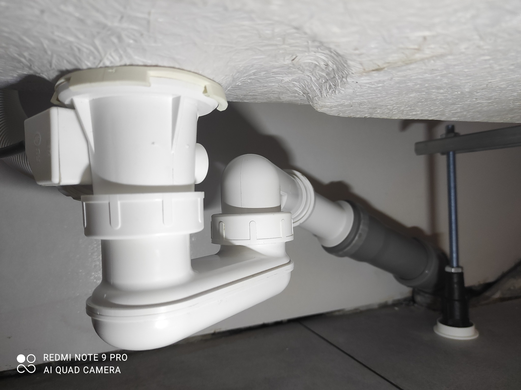 Biały syfon umywalkowy z szarą rurą odpływową, zamontowany pod umywalką w łazience. Widoczne elementy mocujące umywalkę do ściany.