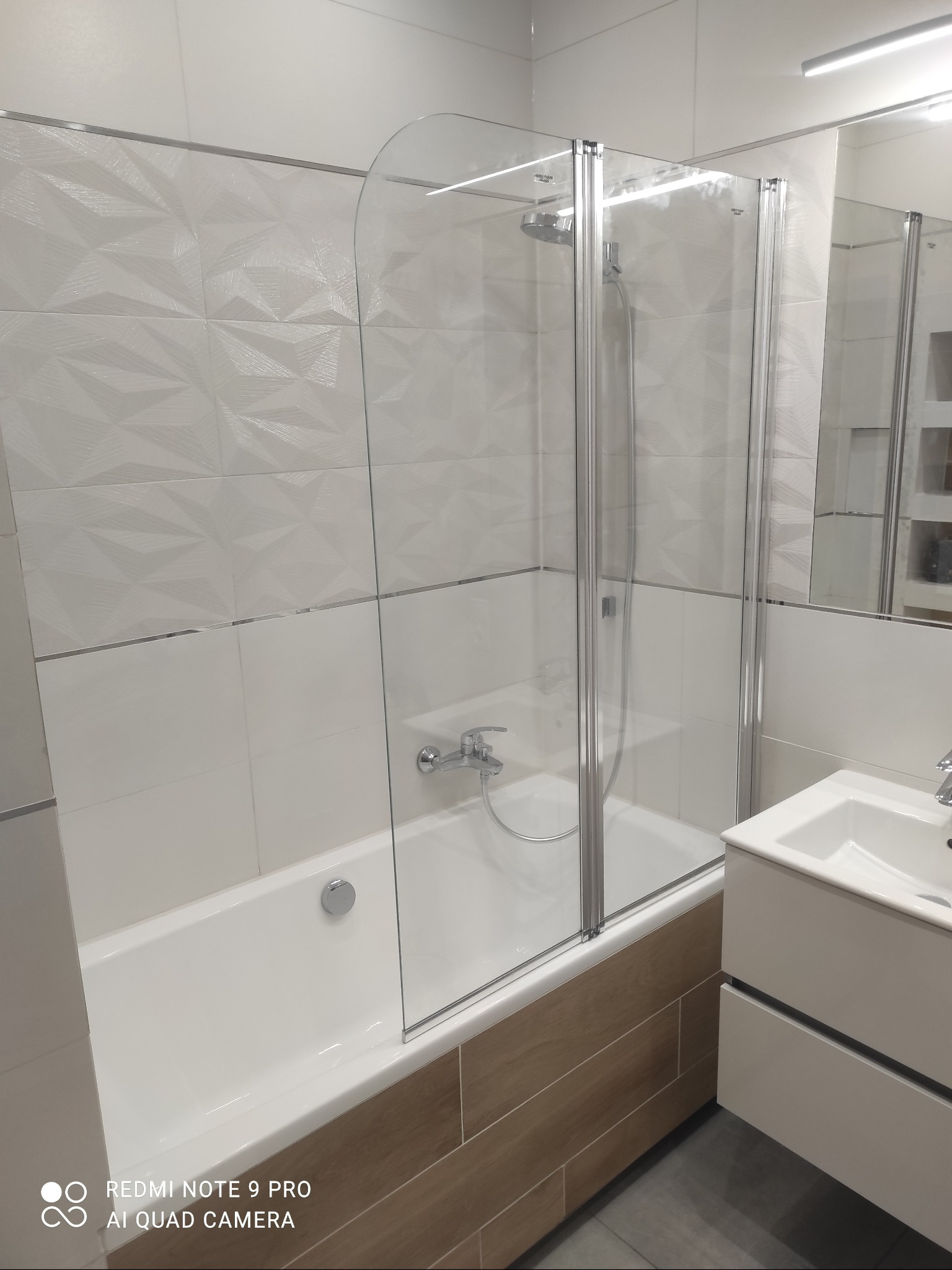 Nowoczesna łazienka z wanną i szklanym parawanem nawannowym, białe płytki z geometrycznym wzorem na ścianie, umywalka z szafką w kolorze złamanej bieli, szare płytki podłogowe.