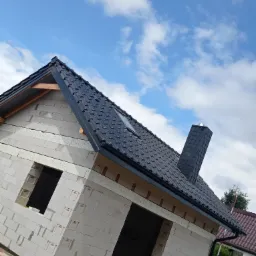 Niedokończony budynek z białego betonu komórkowego z zamontowanym dachem z czarnej dachówki ceramicznej i kominem, widoczne rynny, błękitne niebo z chmurami w tle.