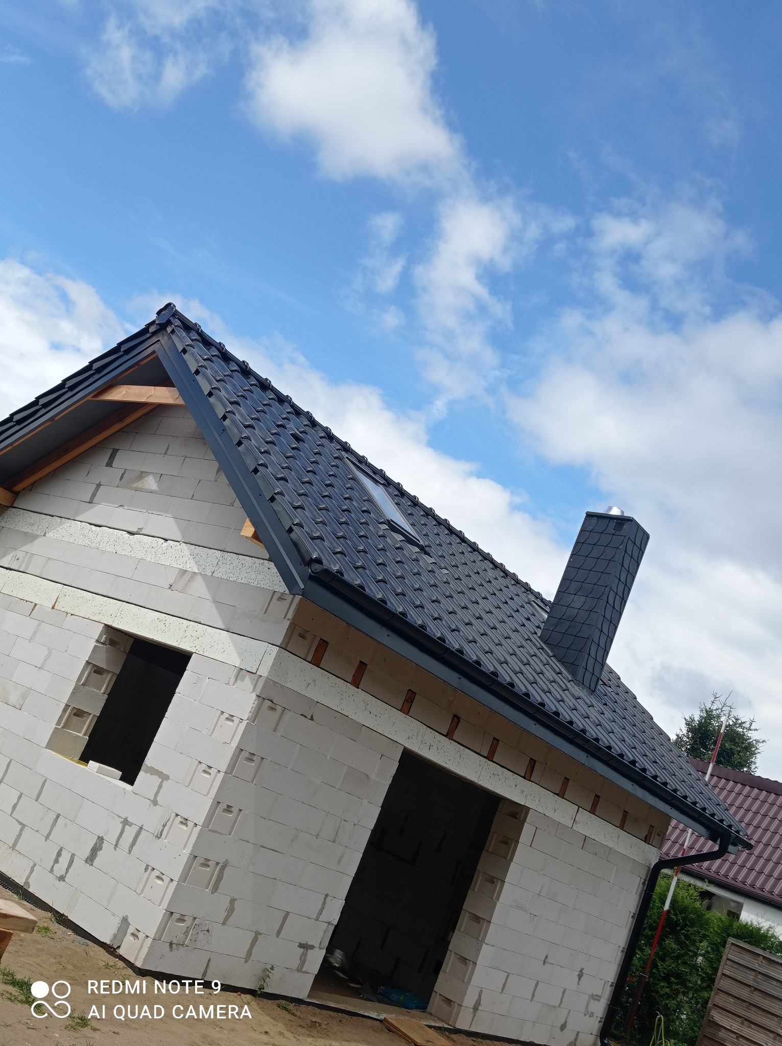 Niedokończony budynek z białego betonu komórkowego z zamontowanym dachem z czarnej dachówki ceramicznej i kominem, widoczne rynny, błękitne niebo z chmurami w tle.