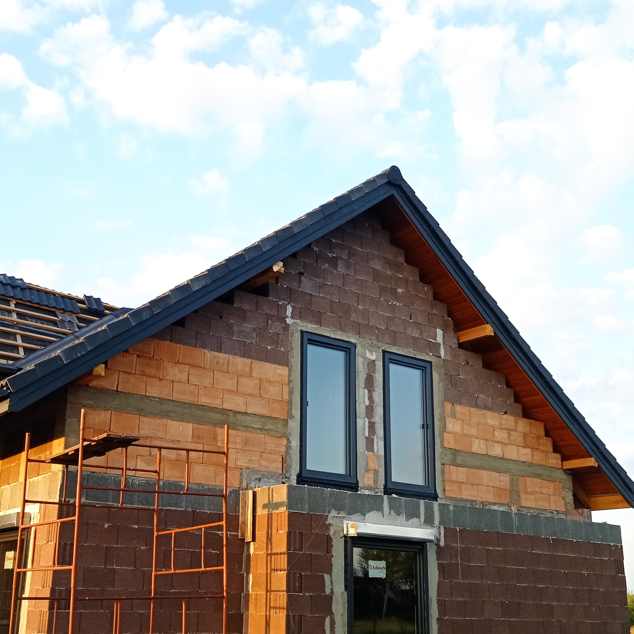 Budowa domu z widoczną konstrukcją dachu, oknami wstawionymi w surowy mur i rusztowaniem w kolorze pomarańczowym, na tle błękitnego nieba z chmurami.