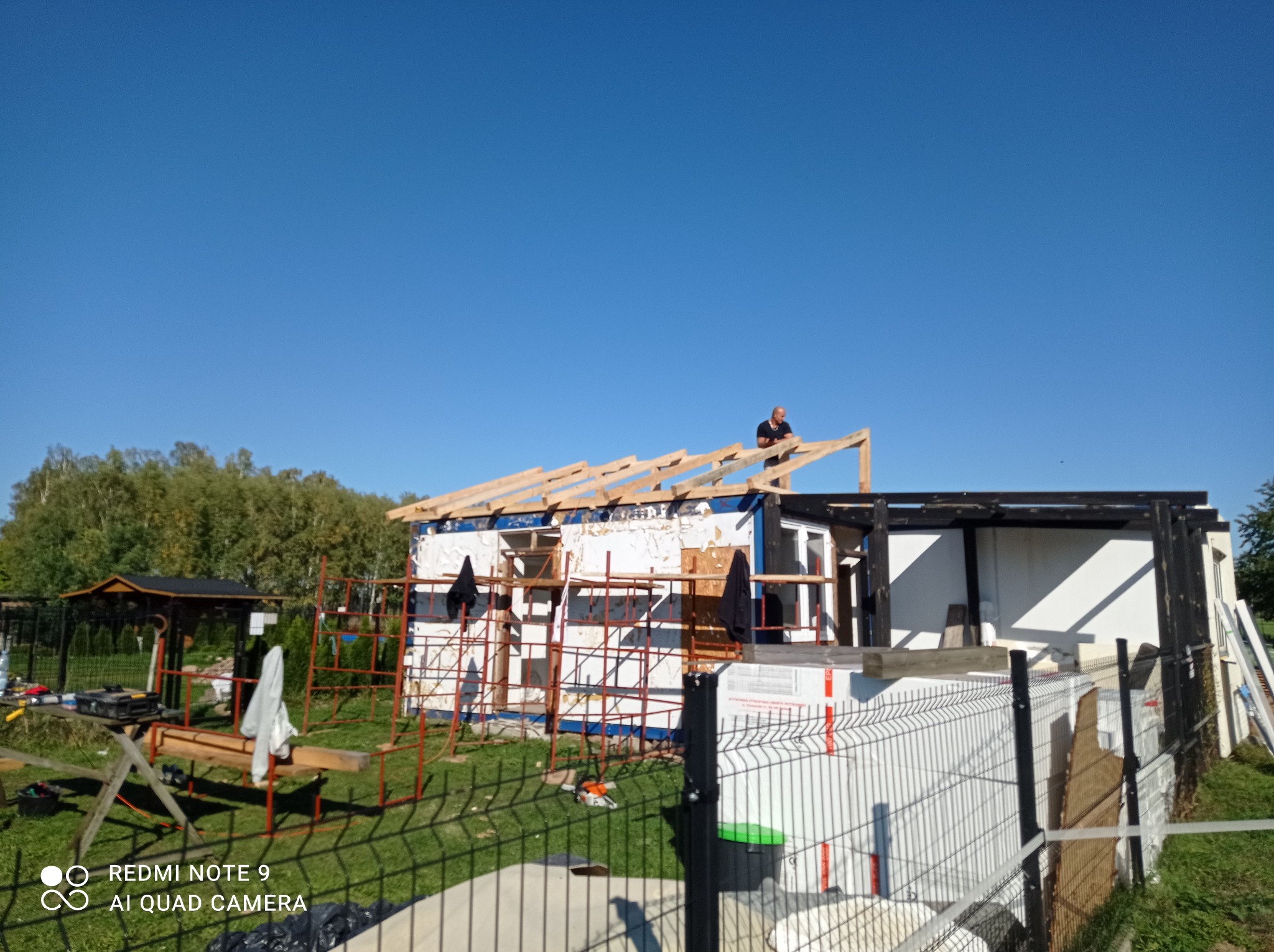 Pracownik na drewnianej konstrukcji dachu budynku modułowego, widoczne rusztowania, folia izolacyjna i narzędzia na trawie, błękitne niebo w tle.