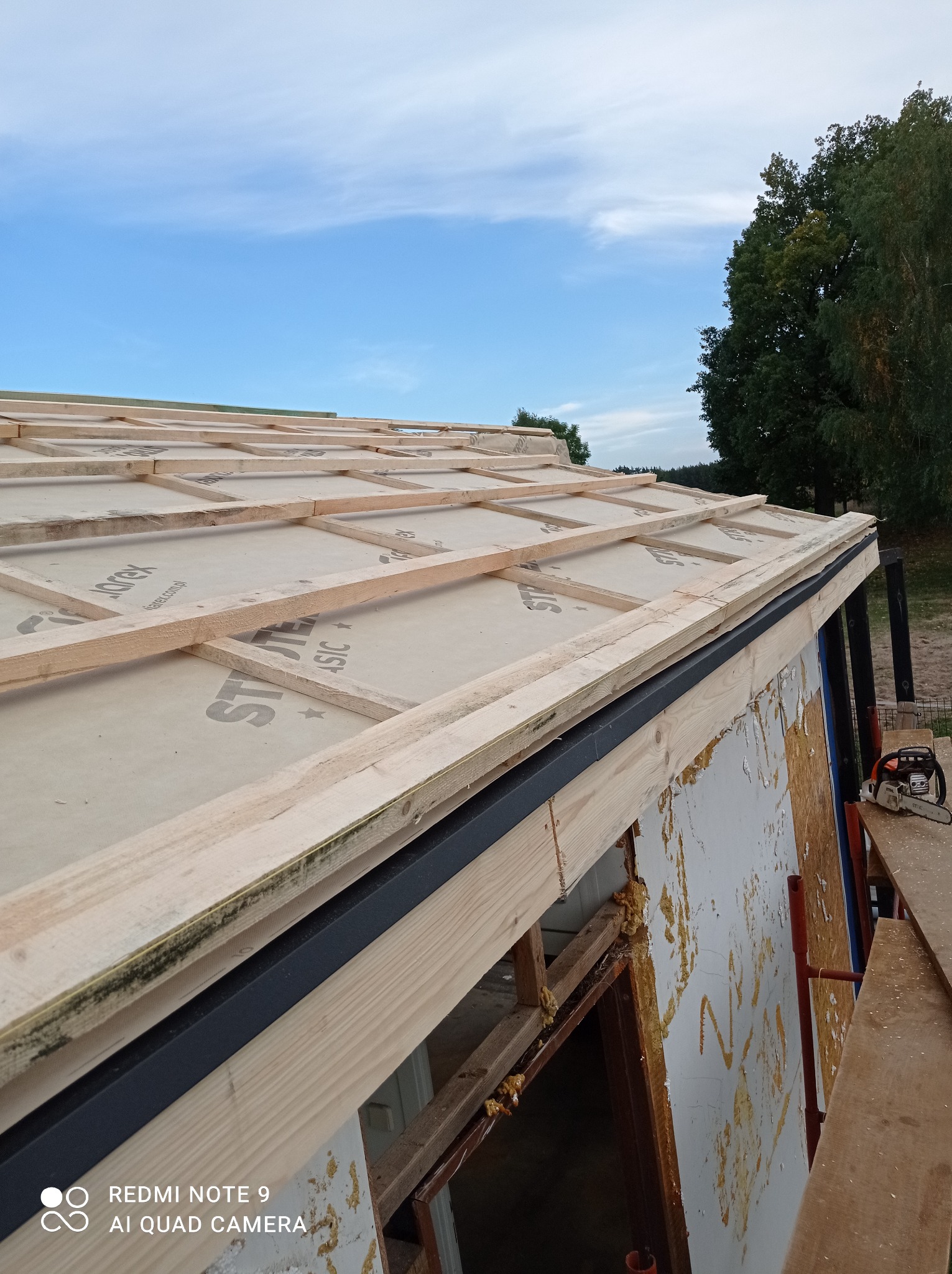 Konstrukcja dachu w trakcie budowy z widoczną membraną dachową, łatami i kontrłatami, narzędzia w tle, widok z boku.