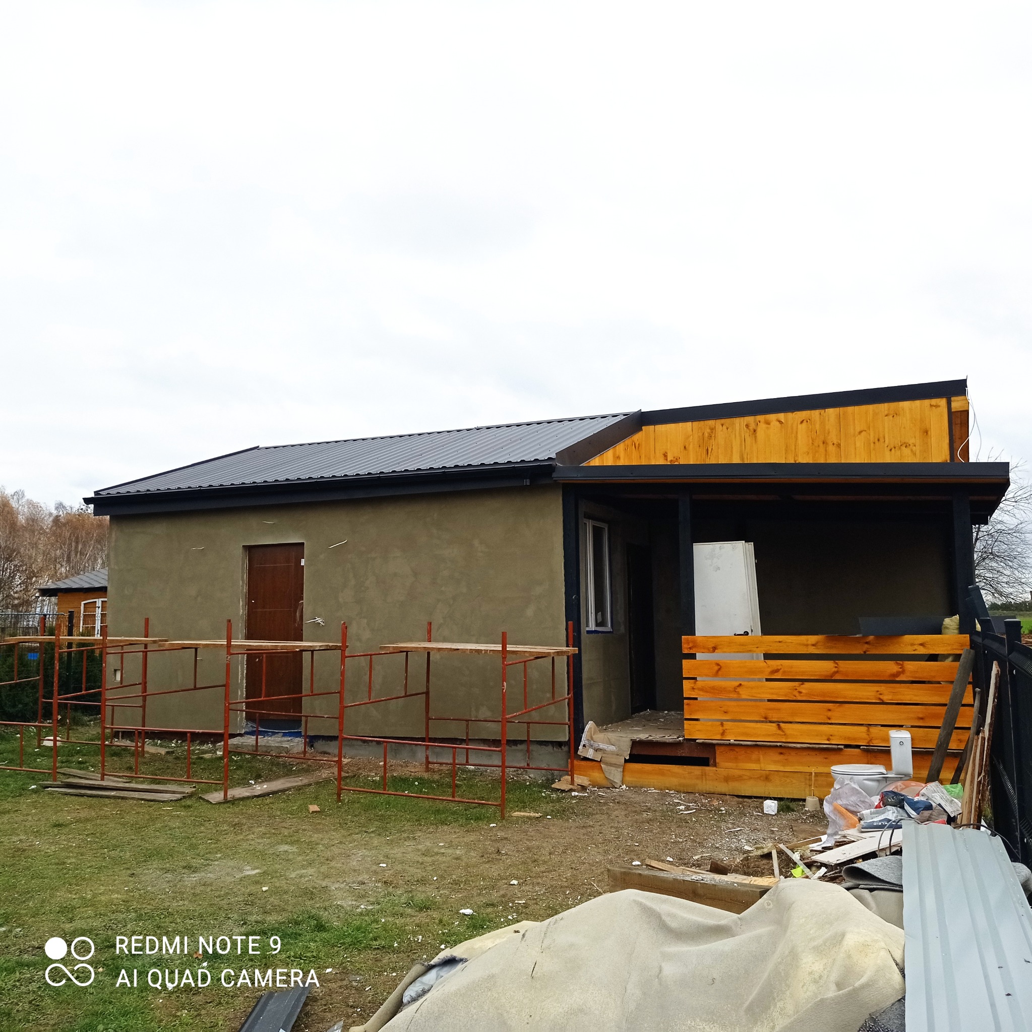 Dom w trakcie budowy z szarym dachem z blachy trapezowej, otynkowaną ścianą, drewnianą werandą i czerwonym rusztowaniem, widok z zewnątrz na posesję z lekkim nieporządkiem.