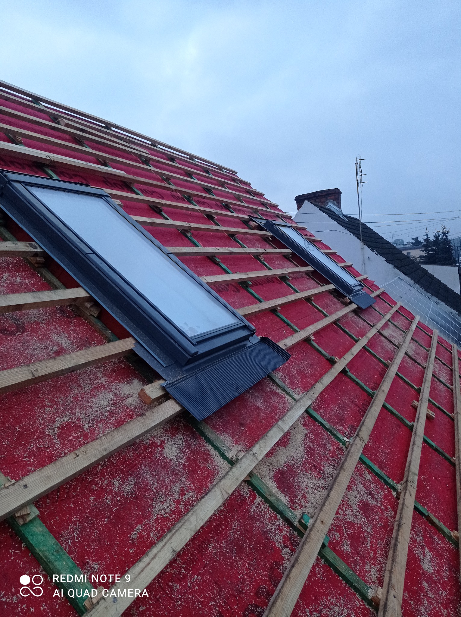 Montaż okien dachowych na czerwonej membranie, widoczna konstrukcja drewniana dachu, w tle dom z instalacją solarną.