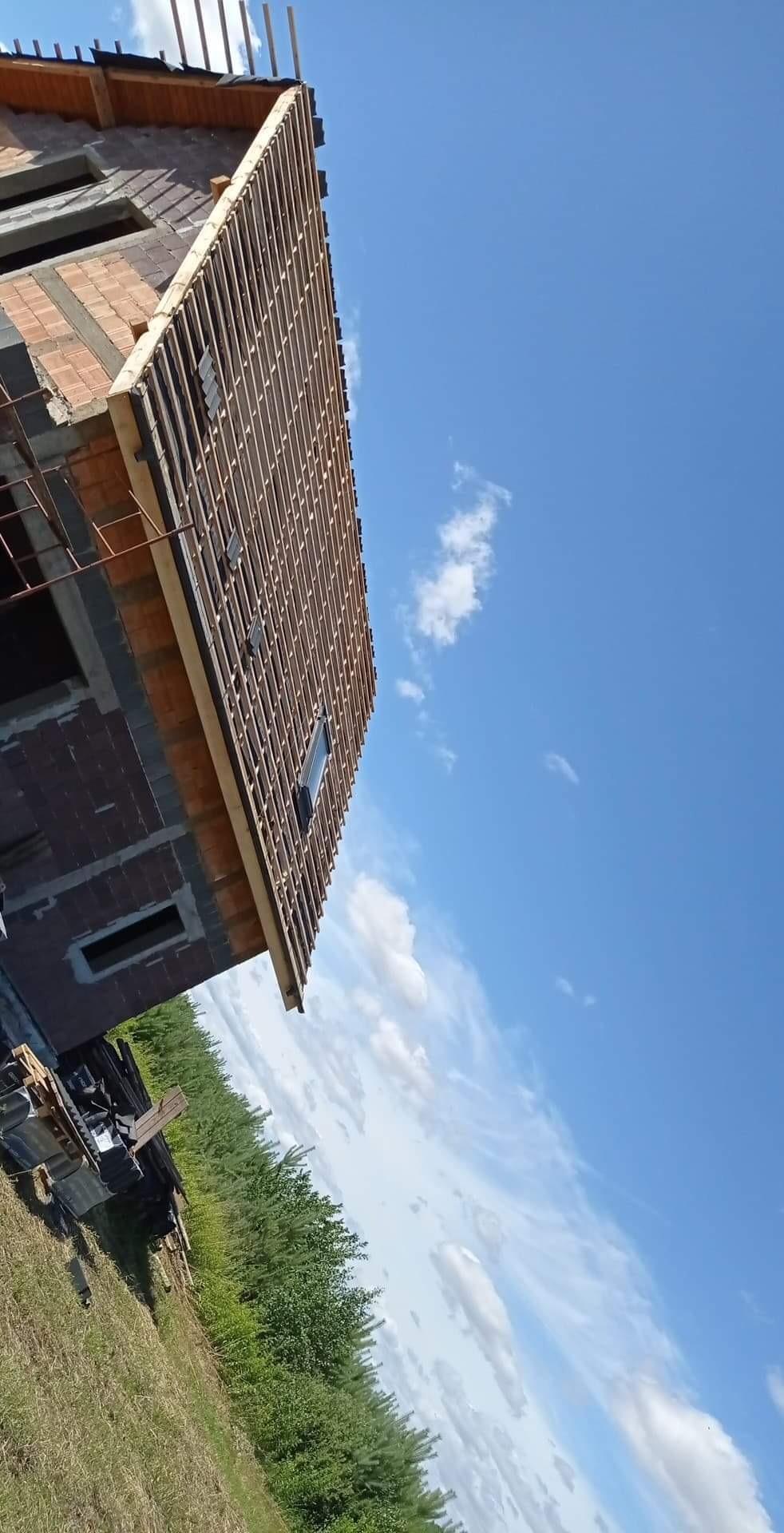 Wznoszenie konstrukcji dachu z drewnianych łat na budynku z czerwonej cegły, widok z dołu na tle błękitnego nieba z chmurami.