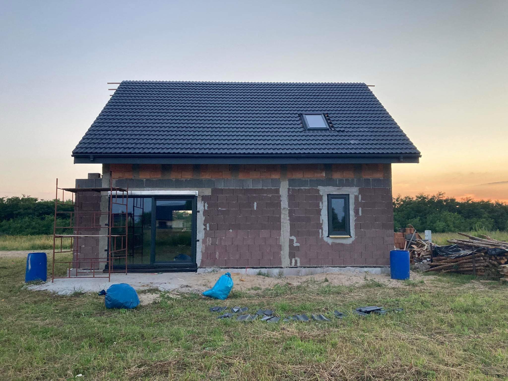 Niedokończony budynek jednorodzinny z nowym, czarnym dachem z dachówką i oknem dachowym, widoczne rusztowanie, okna i fragmenty ścian z cegły, teren wokół budynku zagospodarowany, w tle zachodzące...