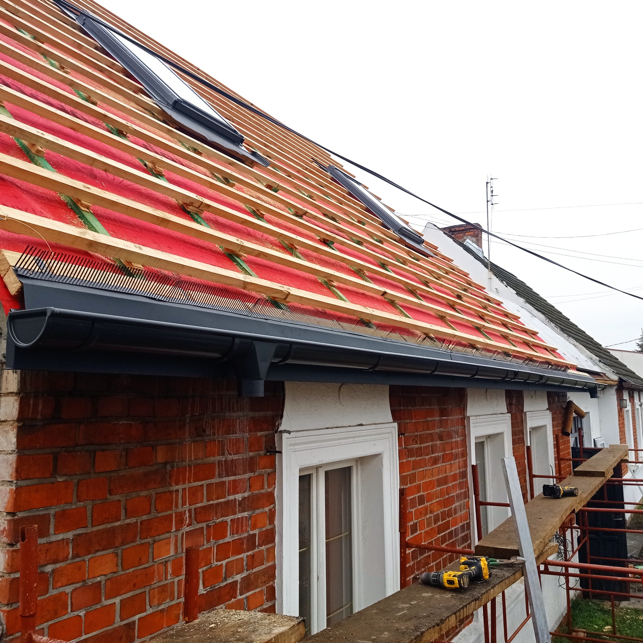 Częściowy widok dachu w trakcie wymiany z zamontowanymi oknami dachowymi, widoczne rynny z zabezpieczeniem przed liśćmi, drewniana konstrukcja dachu z czerwoną membraną, rusztowanie i narzędzia...