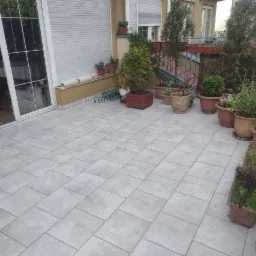 Jasnoszara, kwadratowa terakota na balkonie z roślinami w donicach, widok z wnętrza mieszkania przez otwarte drzwi balkonowe.