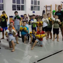 Grupa dzieci i dorosły trener w sali sportowej, pozujący w rękawicach bokserskich, widoczne worki treningowe w tle.