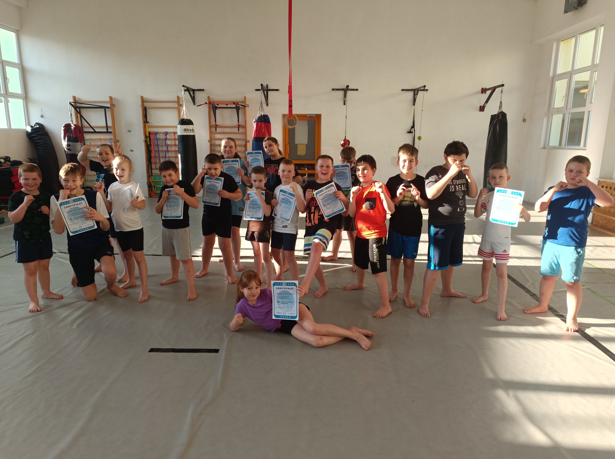 Grupa dzieci, boso, z certyfikatami w dłoniach, po treningu na sali sportowej z workiem bokserskim i drabinkami gimnastycznymi w tle.