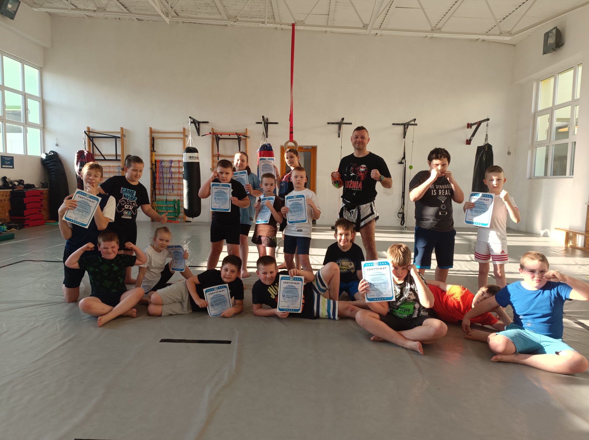Grupa dzieci i trener sportów walki w sali treningowej, dzieci siedzą na macie i trzymają certyfikaty ukończenia kursu, w tle worki treningowe i drabinki.