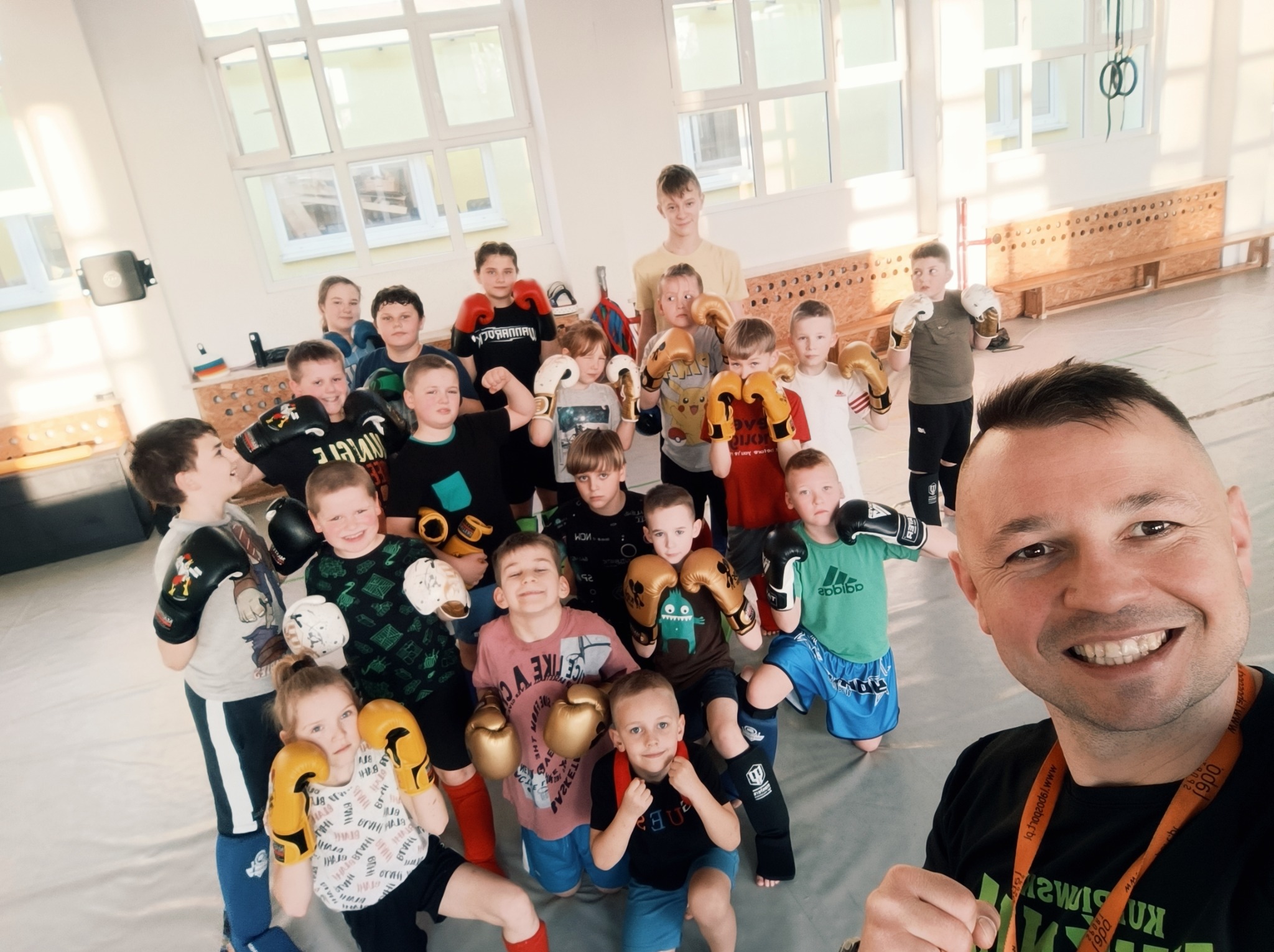 Grupa dzieci w różnym wieku, ubrana w rękawice bokserskie, pozuje do selfie z uśmiechniętym trenerem w sali sportowej z oknami i drabinkami gimnastycznymi w tle. Dzieci mają różne kolory rękawic...