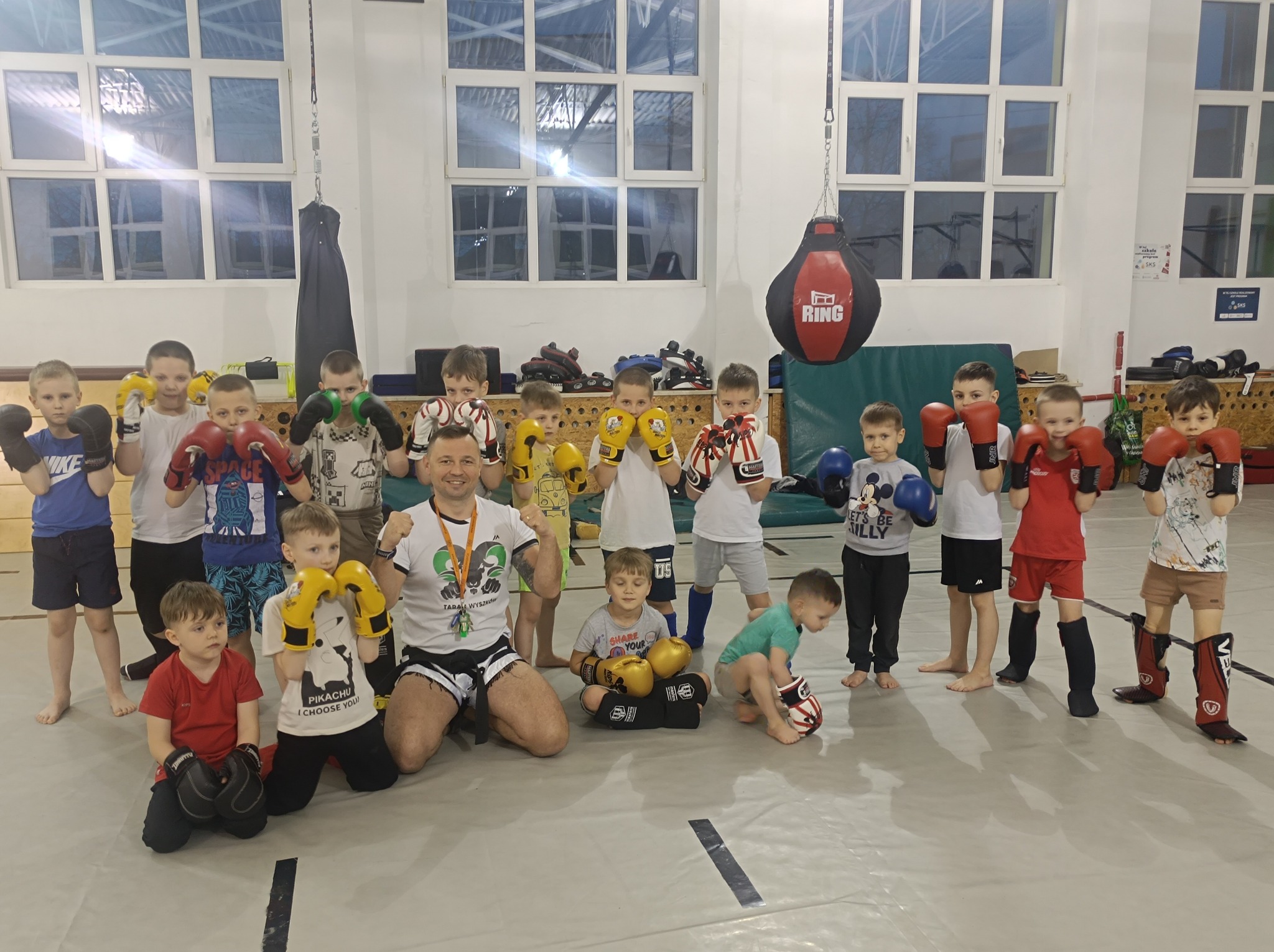 Grupa dzieci w różnym wieku w rękawicach bokserskich i ochraniaczach, pozująca z trenerem na sali treningowej ze sprzętem do sportów walki w tle.