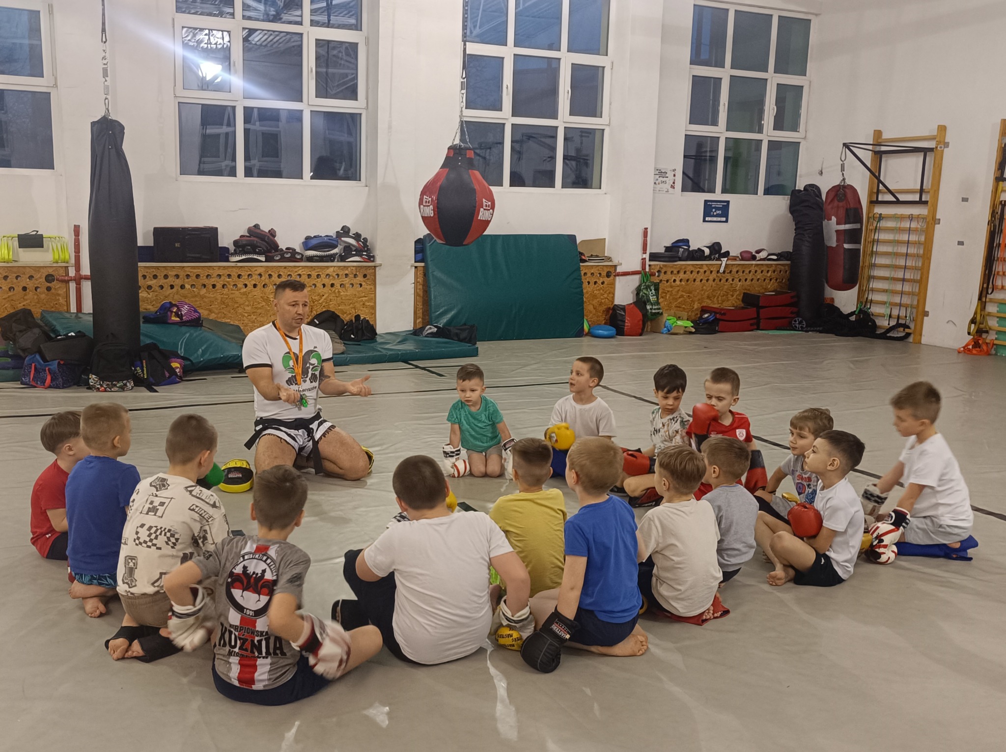 Instruktor sportów walki prowadzi zajęcia dla grupy małych dzieci w sali treningowej z workiem bokserskim i drabinkami gimnastycznymi w tle. Dzieci siedzą w kręgu, część ma na sobie rękawice...