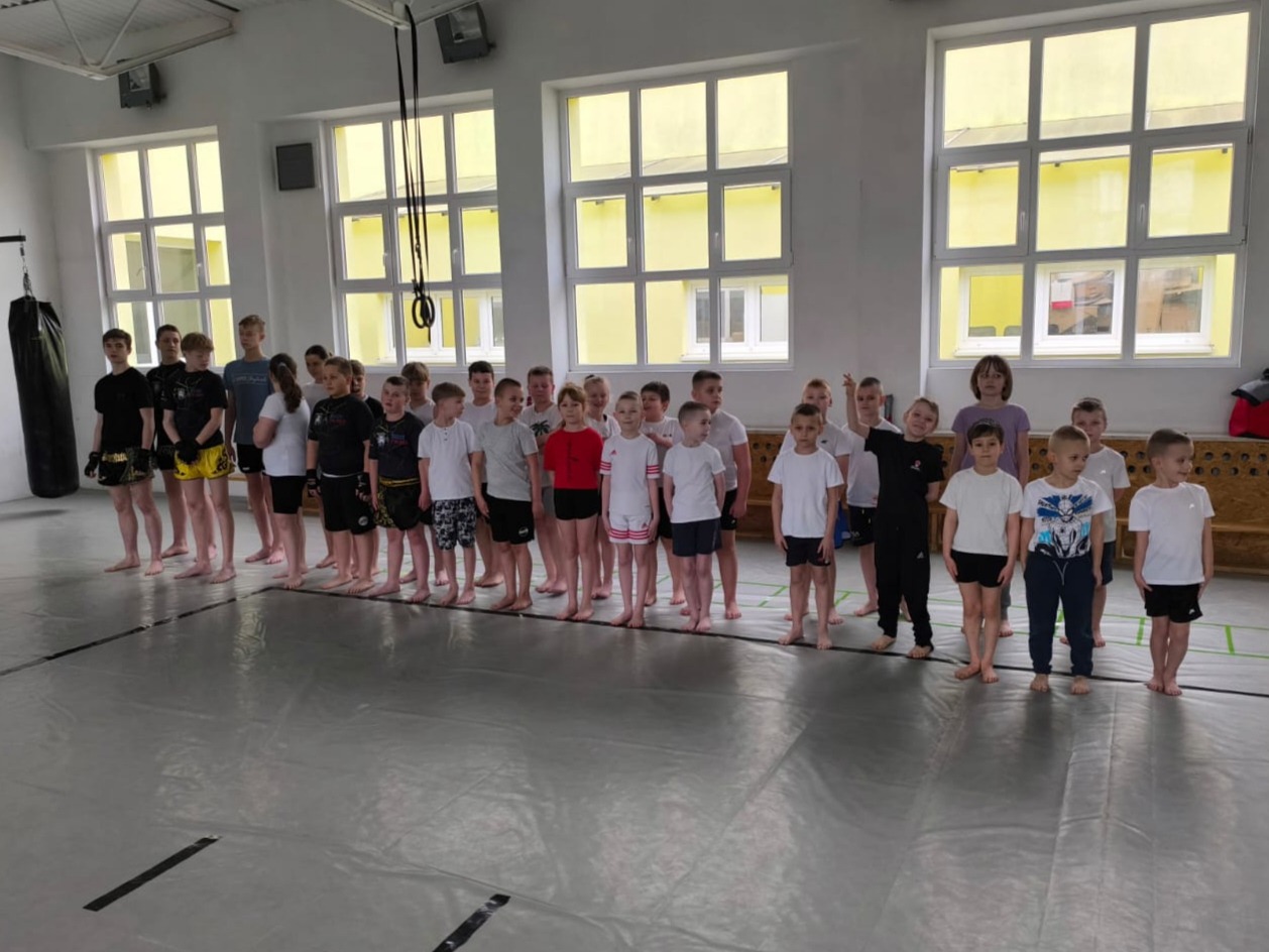 Grupa dzieci i młodzieży ustawiona w rzędzie na sali treningowej w Wyszkowie, prawdopodobnie po zajęciach sportów walki; worek treningowy widoczny w tle po lewej stronie.