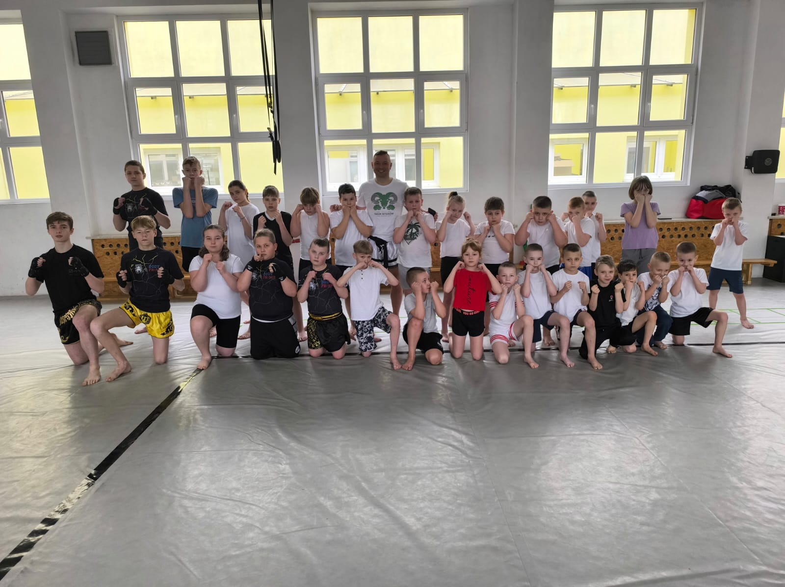Grupa dzieci i trener w sali gimnastycznej, ustawieni w rzędach, przygotowują się do treningu sportów walki, ustawiając gardę.