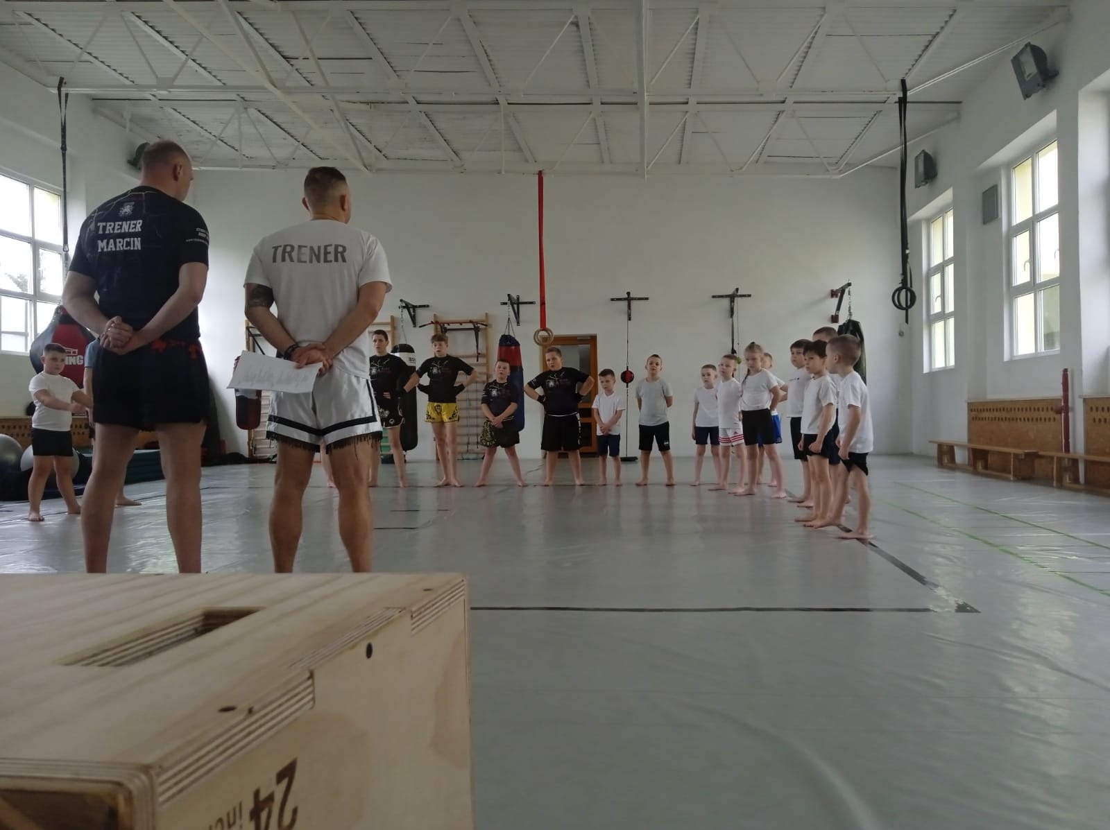 Trening sportów walki dla dzieci i młodzieży w sali gimnastycznej, prowadzony przez dwóch trenerów, widoczna skrzynia do ćwiczeń.