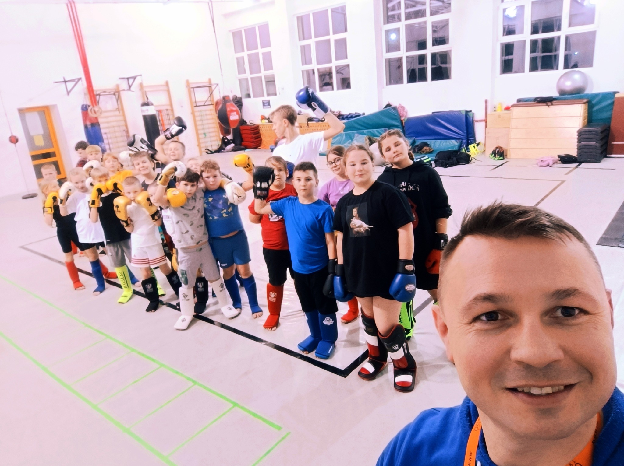 Grupa dzieci w różnokolorowych strojach i rękawicach bokserskich ustawiona w rzędzie w sali gimnastycznej, z uśmiechniętym mężczyzną robiącym selfie na pierwszym planie.