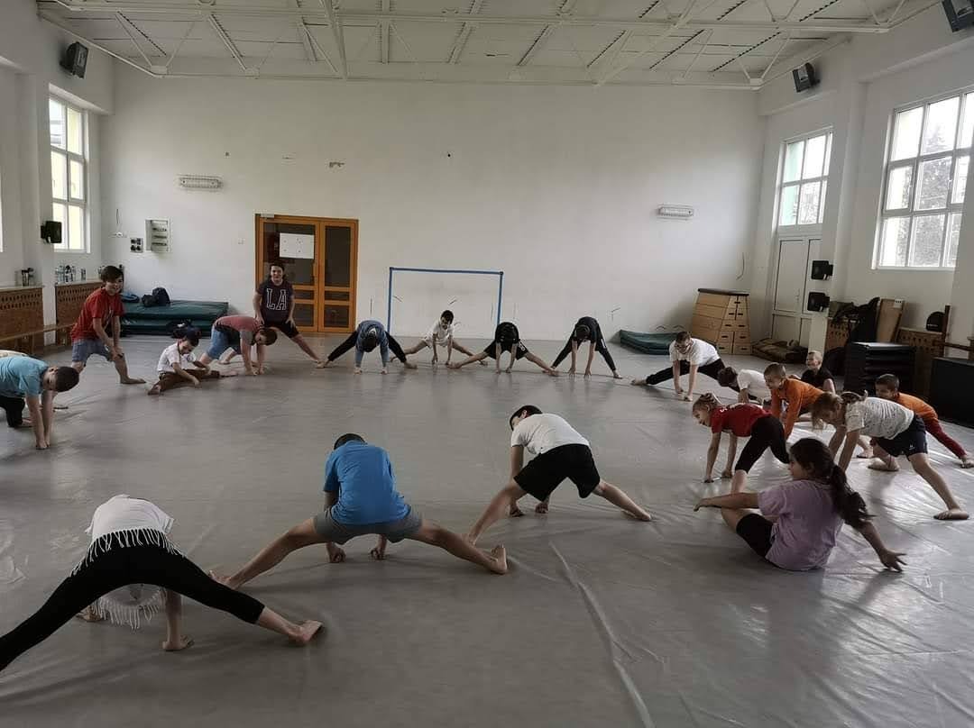 Grupa dzieci rozciąga się na podłodze w sali gimnastycznej, wykonując szpagat pod nadzorem trenera, widoczne okna i wyposażenie sali.