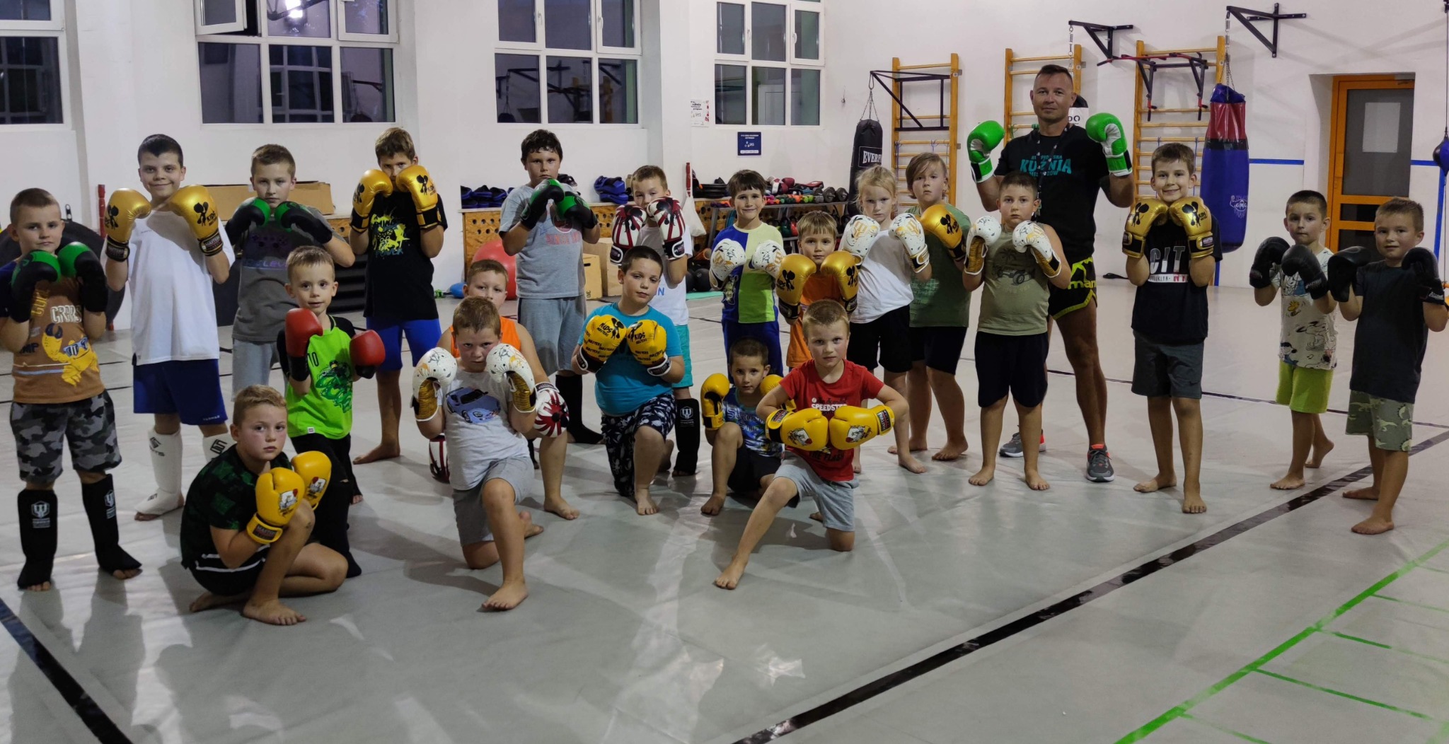 Grupa dzieci i dorosły trener w sali sportowej, pozujący w rękawicach bokserskich, widoczne worki treningowe w tle.