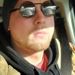 Mężczyzna w samochodzie, w czapce z logo Red Bulla i okularach przeciwsłonecznych odbijających drogę, ubrany w bluzę i kurtkę, zapięty pasem bezpieczeństwa.