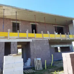 Surowy stan dwukondygnacyjnego domu z betonu komórkowego z widocznymi podporami stropu i żółtym deskowaniem, paleta z białymi bloczkami oraz stos czerwonych cegieł na pierwszym planie.