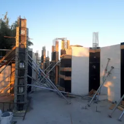 Wznoszenie ścian z bloczków betonowych z użyciem szalunków i podpór teleskopowych, widoczne zbrojenia pionowe i zachodzące słońce.
