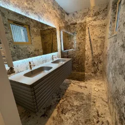 Nowoczesna łazienka z podwójną umywalką na szafce z lamelami, lustrem z podświetleniem LED i ścianami wykończonymi w stylu imitującym kamień.