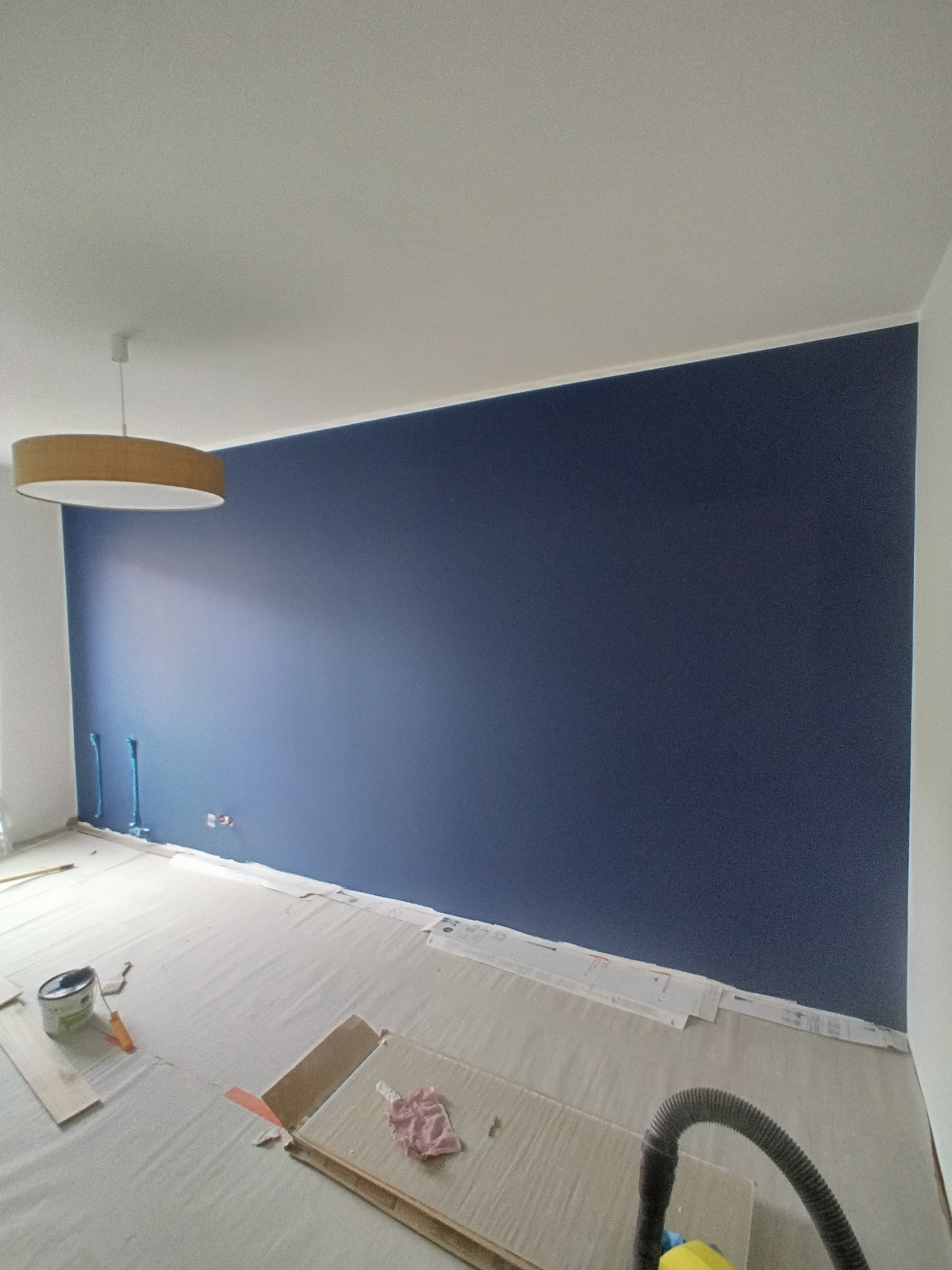 Pomieszczenie w trakcie malowania: ściana pomalowana na ciemny granat, zabezpieczona podłoga, widoczna puszka z farbą i narzędzia malarskie.