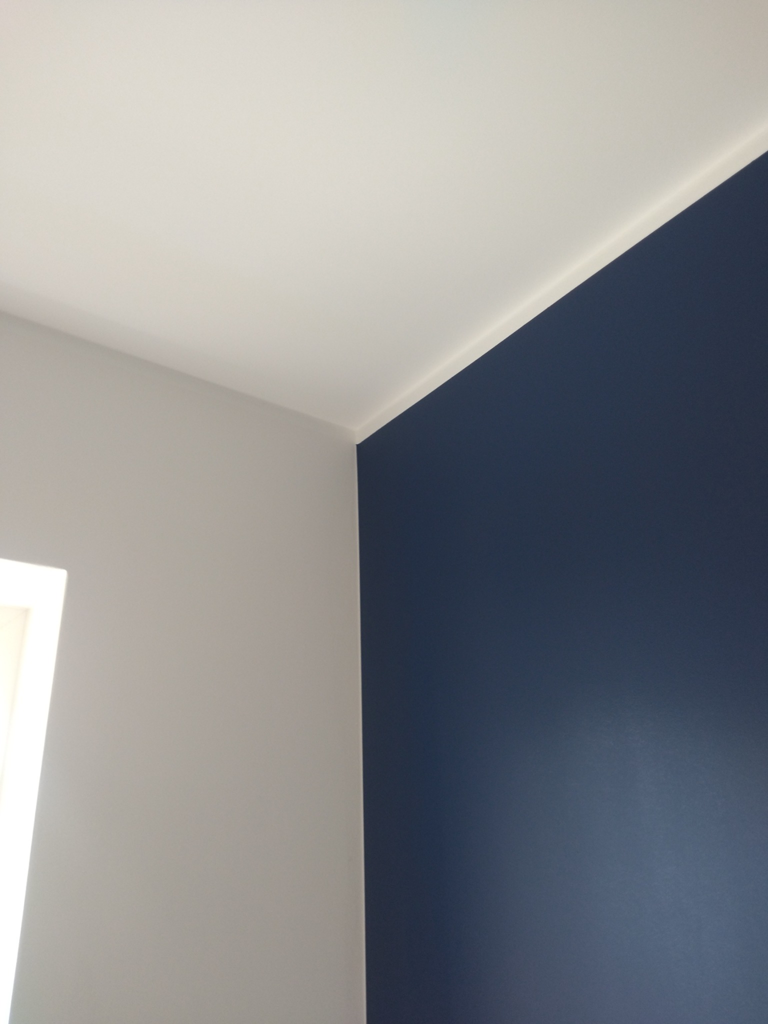 Fragment pomalowanego wnętrza: granatowa ściana z listwą przypodłogową, szara ściana i biały sufit, widoczny fragment okna.