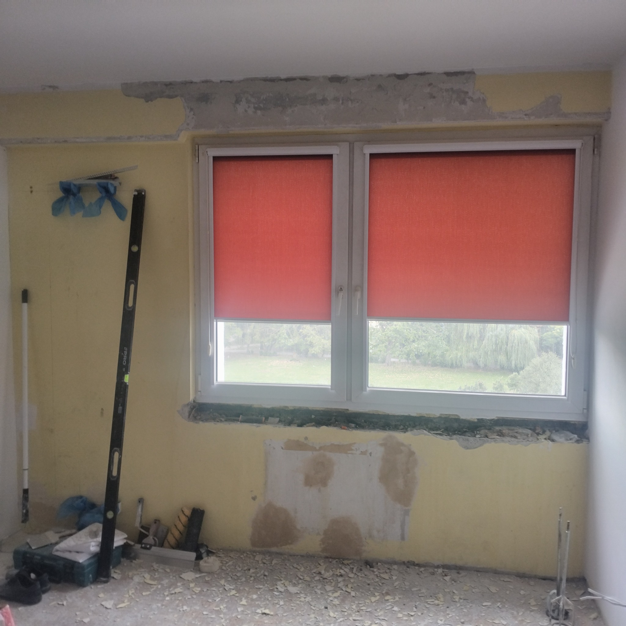 Pokój w trakcie prac przygotowawczych: częściowo usunięta stara farba, ślady po szpachlowaniu, czerwone rolety w oknie, poziomica oparta o ścianę, narzędzia na podłodze.