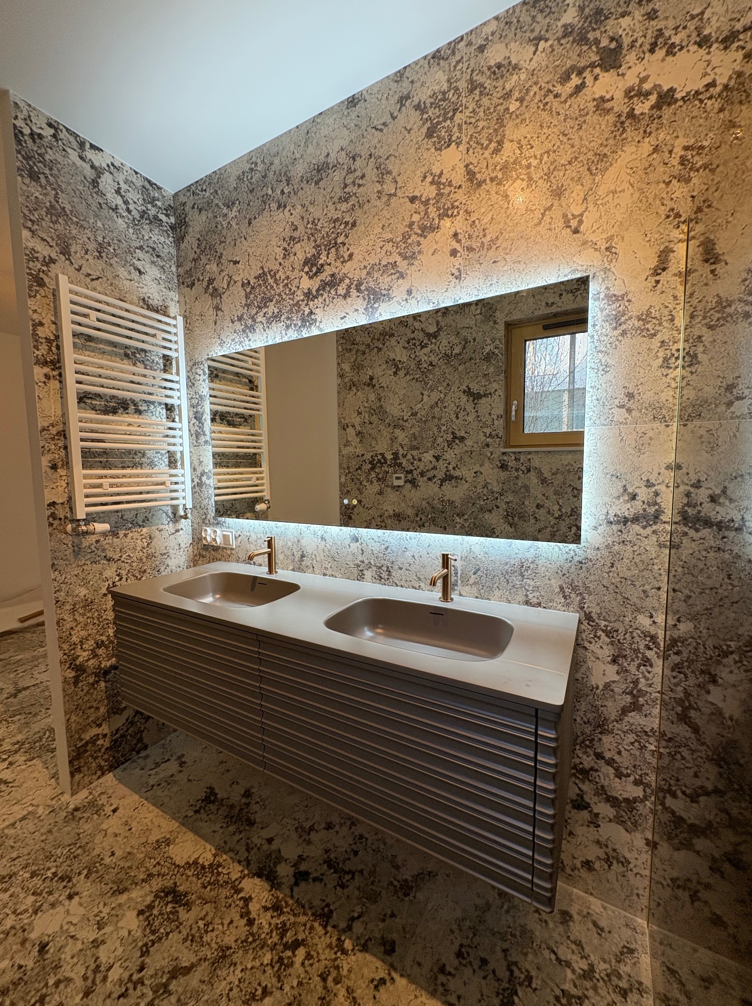 Nowoczesna łazienka z dwoma umywalkami w szarej szafce z lamelami, podświetlanym lustrem i grzejnikiem drabinkowym na ścianie z efektem dekoracyjnej faktury.