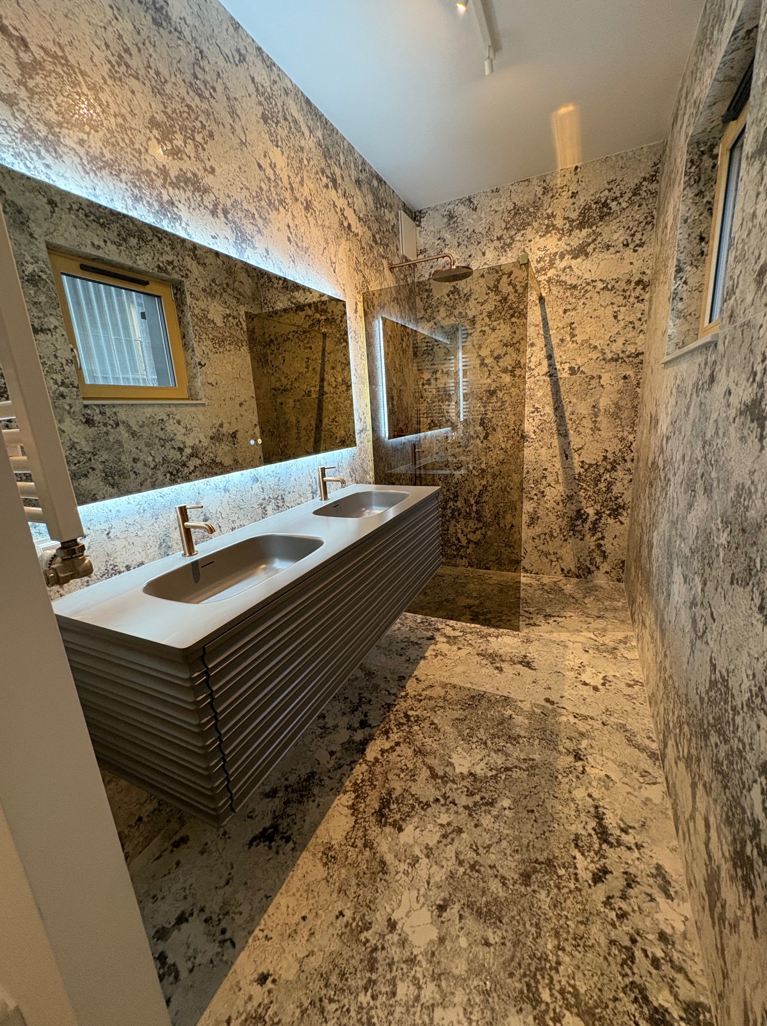 Nowoczesna łazienka z podwójną umywalką na szafce z lamelami, lustrem z podświetleniem LED i ścianami wykończonymi w stylu imitującym kamień.