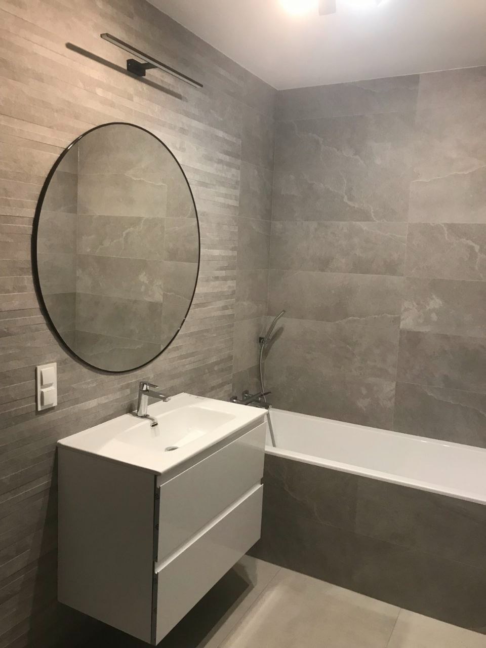 Nowoczesna łazienka z szarymi płytkami imitującymi kamień, okrągłym lustrem z czarną ramą nad białą szafką z umywalką i wanną.