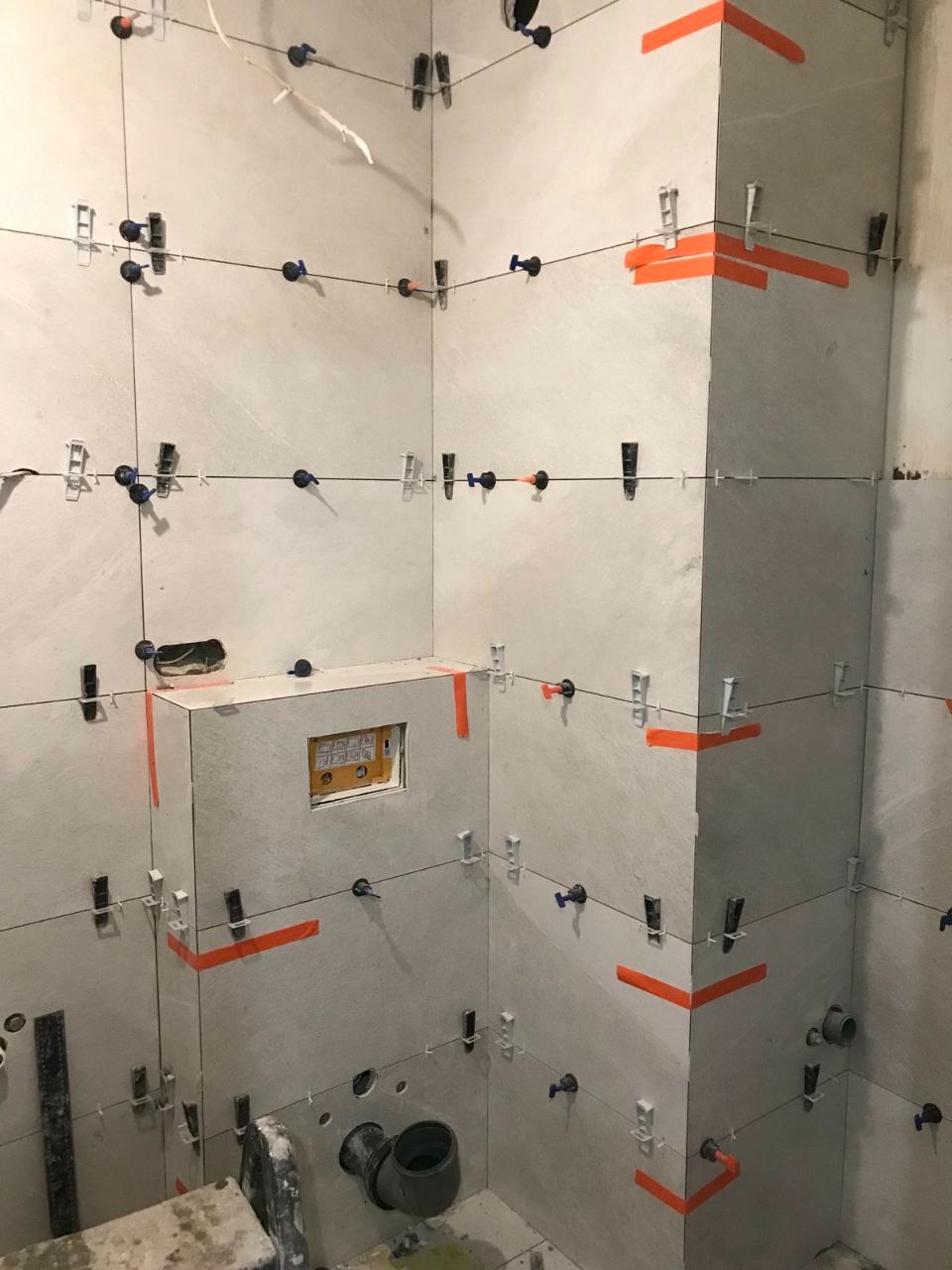 W trakcie układania płytek ściennych w rogu łazienki, widoczne klipsy poziomujące i pomarańczowa taśma maskująca, przygotowane otwory na instalacje sanitarne.