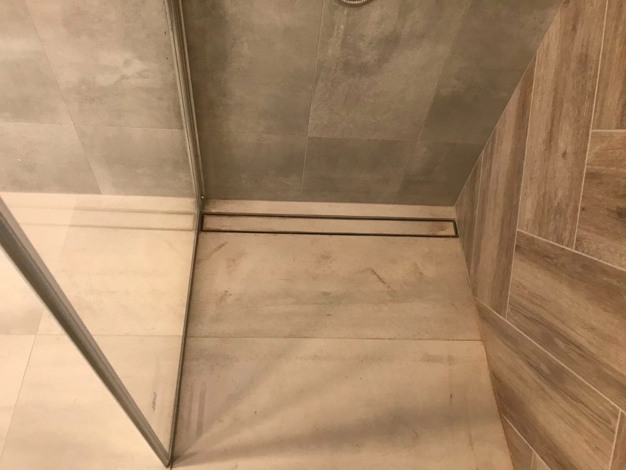 Wykończona kabina prysznicowa z odpływem liniowym, wyłożona płytkami w odcieniach beżu i szarości, z widocznym fragmentem szklanej ściany.