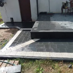 Nowo ułożony taras z ciemnej kostki brukowej z jasnymi obrzeżami, prowadzący do drzwi budynku, widoczne narzędzia i węże ogrodowe.