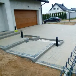 Nowo ułożona, jasnoszara nawierzchnia z kostki brukowej przed domem z garażem i brązową bramą, z elementami dekoracyjnymi obramowanymi ciemniejszą kostką, widoczne lampki oświetleniowe i częściowo...