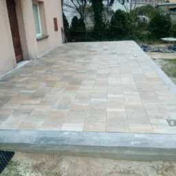 Nowo ułożony taras z jasnych, prostokątnych płyt na betonowej wylewce, widok z boku domu w pochmurny dzień.