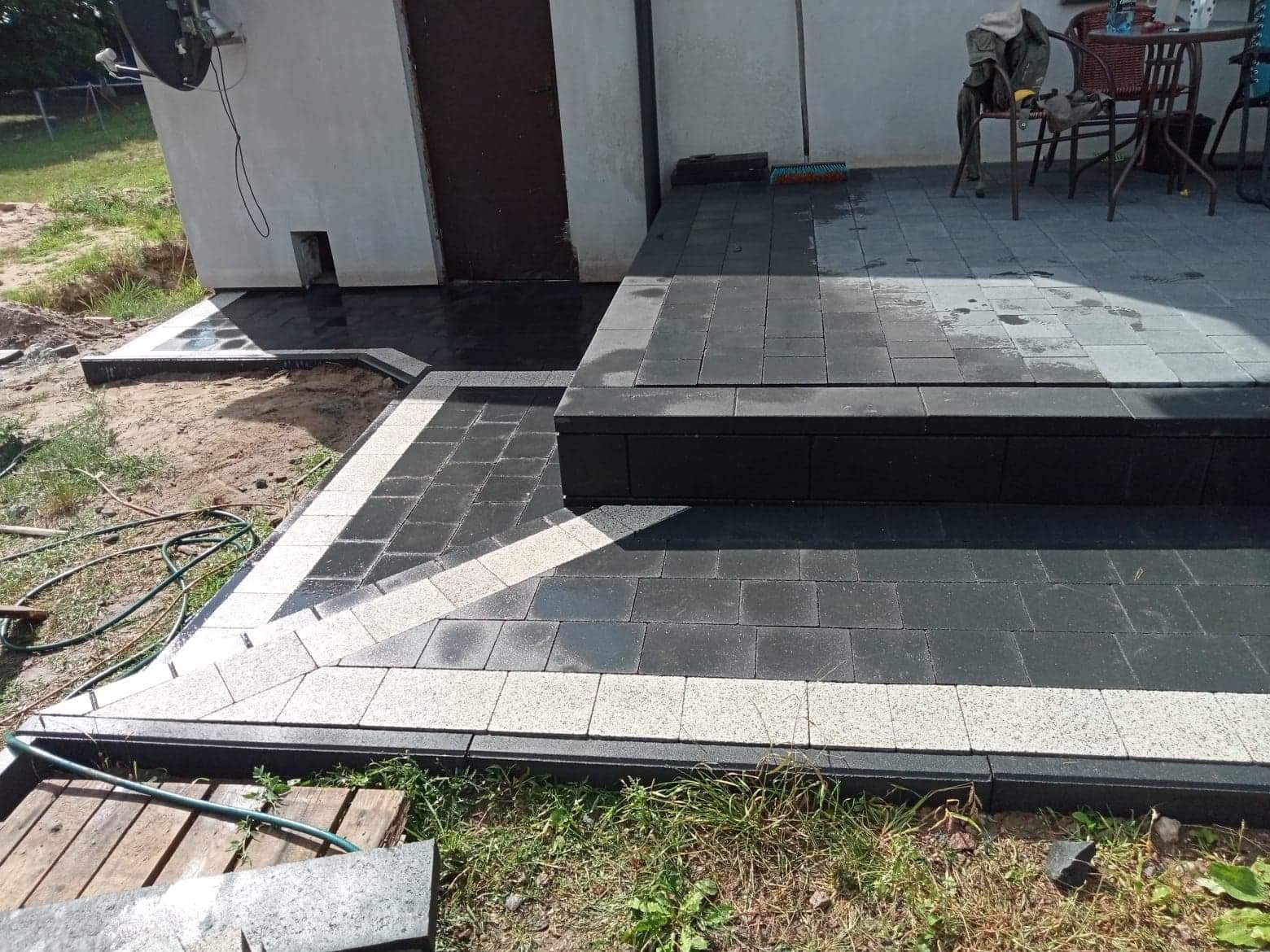 Nowo ułożony taras z ciemnej kostki brukowej z jasnymi obrzeżami, prowadzący do drzwi budynku, widoczne narzędzia i węże ogrodowe.