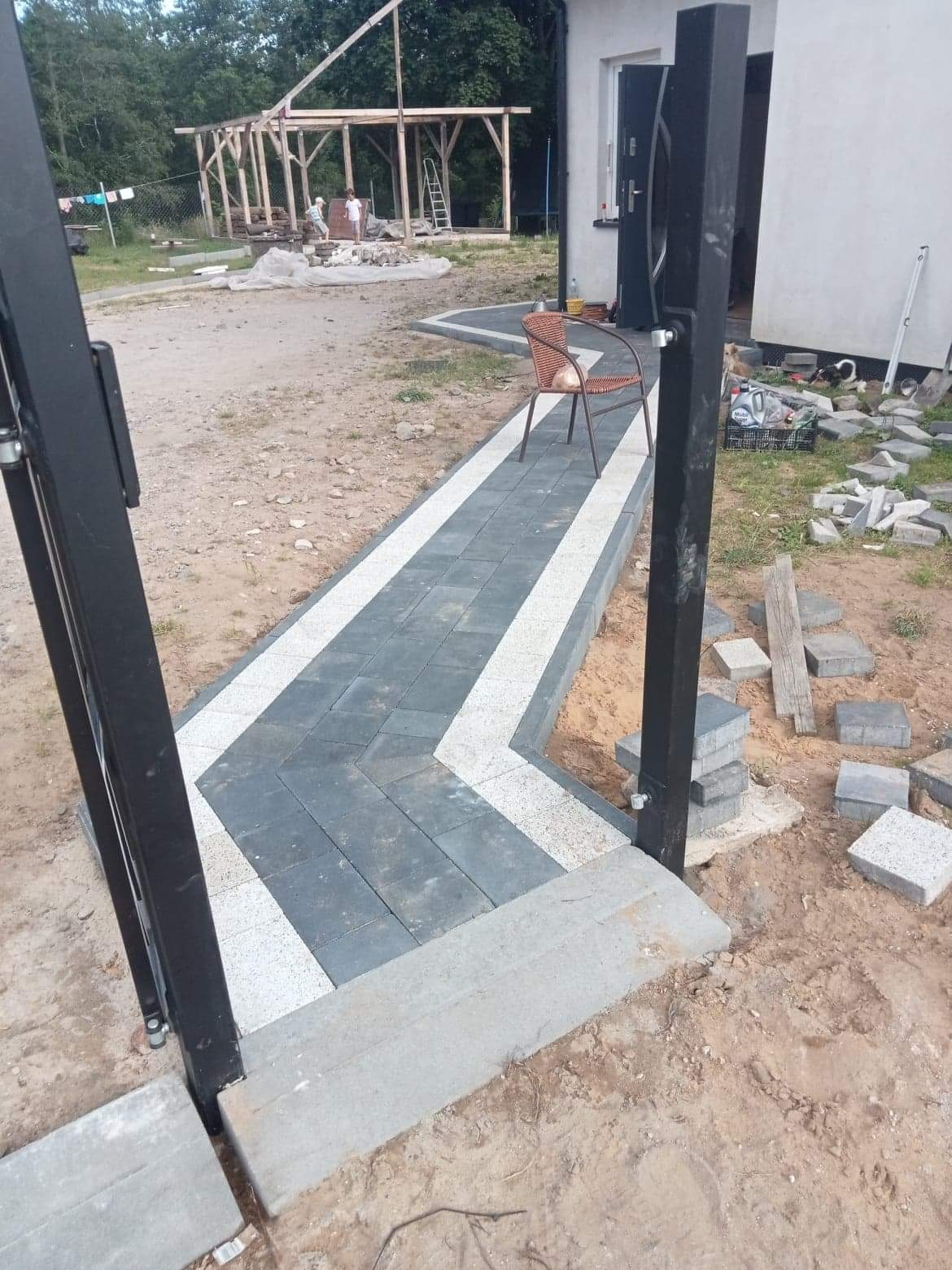 Ścieżka z kostki brukowej w kolorach szarości i bieli, ułożona we wzór zygzak, prowadząca od furtki do domu. W tle widoczna konstrukcja drewniana i materiały budowlane.