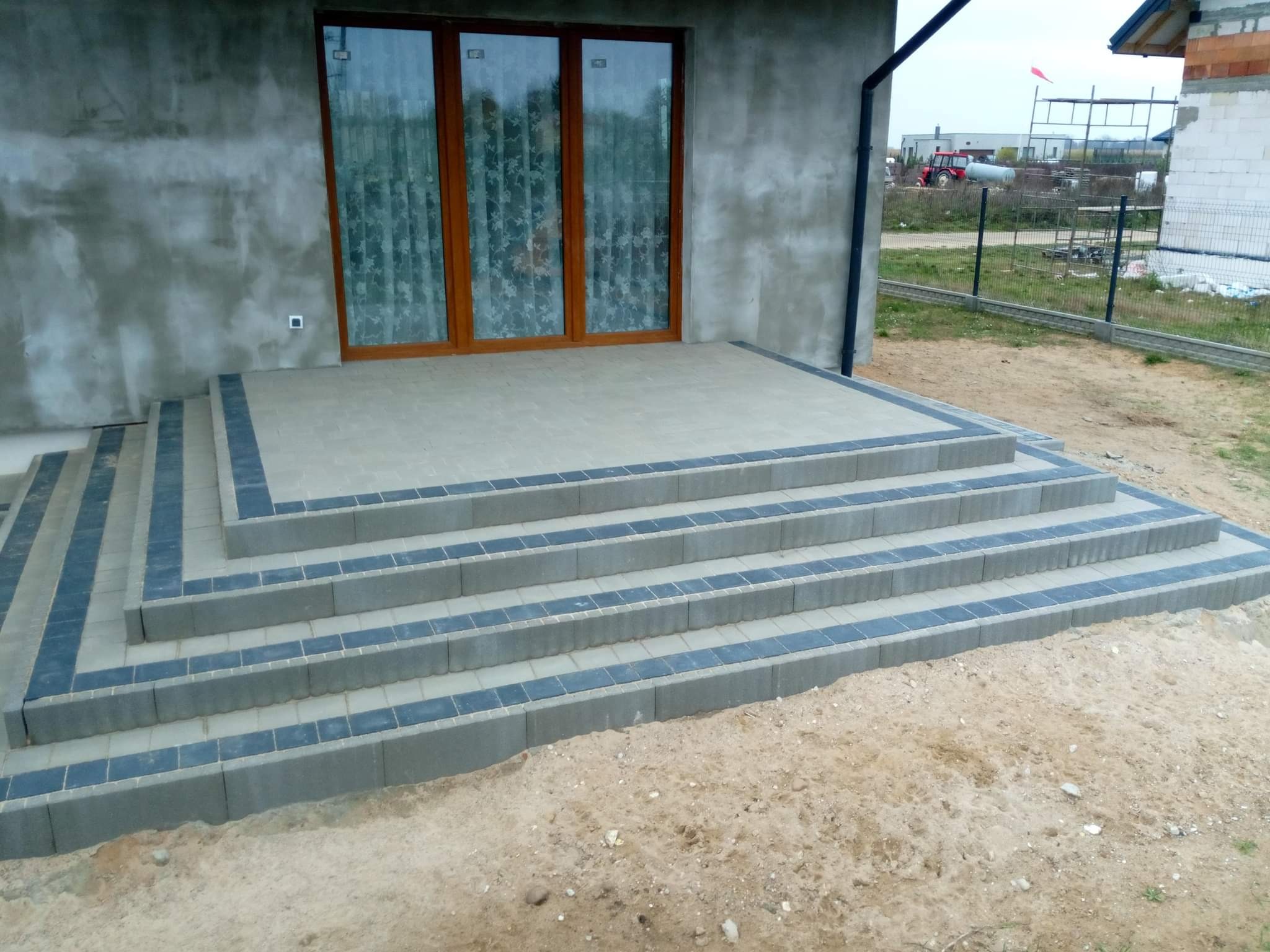 Schody wejściowe wyłożone szarą kostką brukową z ciemnymi obwódkami, prowadzące do domu w stanie surowym z drewnianą stolarką okienną.