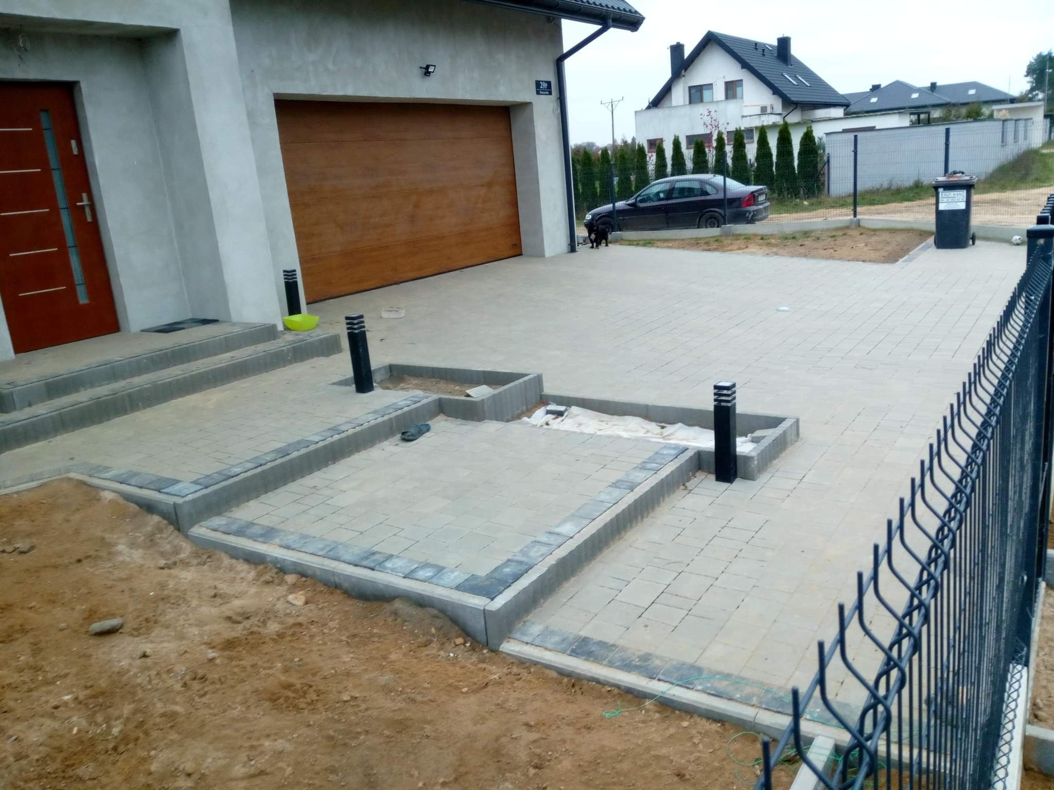 Nowo ułożona, jasnoszara nawierzchnia z kostki brukowej przed domem z garażem i brązową bramą, z elementami dekoracyjnymi obramowanymi ciemniejszą kostką, widoczne lampki oświetleniowe i częściowo...