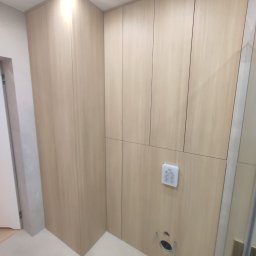 Rafał Grządziel RAFMEB Usługi Stolarskie - Jasne, drewniane szafy wnękowe w łazience z widocznym panelem sterowania i otworem wentylacyjnym w dolnej części.