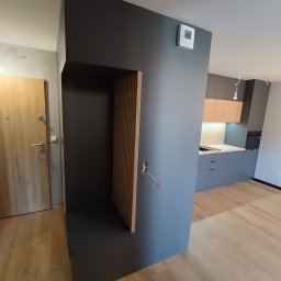 Rafał Grządziel RAFMEB Usługi Stolarskie - Nowoczesne wnętrze z antracytową ścianą działową, w której wbudowana jest szafa z lamelami; w tle widoczna szara zabudowa kuchenna z drewnianymi szafkami.