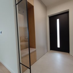 Rafał Grządziel RAFMEB Usługi Stolarskie - Nowoczesny przedpokój z zabudowaną szafą wnękową z siedziskiem i czarnymi, metalowymi elementami wykończeniowymi, widok na czarne drzwi wejściowe z przeszkleniem.