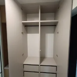 Otwarta szafa wnękowa z szufladami i półkami, widoczne zawiasy na drzwiach, jasne wykończenie.