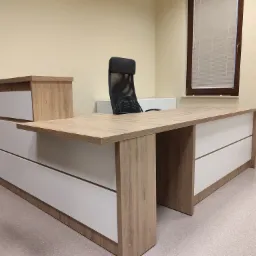 Biurko narożne z blatem imitującym jasne drewno i białymi frontami szuflad, z krzesłem biurowym na tle okna z żaluzjami.